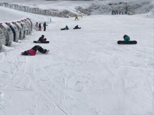 Schneebedeckte Piste mit mehreren Menschen auf Snowboards und einige liegen im Schnee.