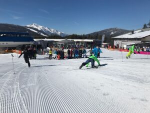 Skifahrer und Zuschauer auf einer Skipiste bei sonnigem Wetter. Berge im Hintergrund.
