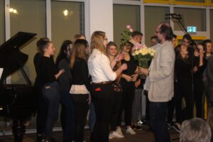Aufführung mit Schülern, die Blumen von einem Mann im Anzug erhalten. Klavier im Hintergrund.
