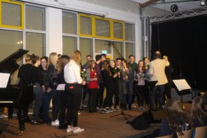 Schüler singen auf der Bühne mit Blumen in der Hand, Klavier im Hintergrund.