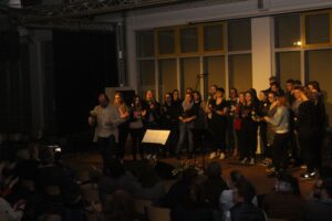 Chorauftritt in einem Veranstaltungsraum mit Publikum und Musikern.