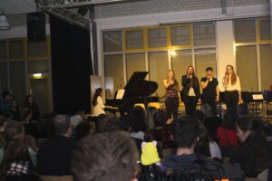 Schüler singen vor Publikum, während eine Schülerin am Klavier spielt.