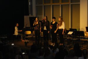 Schülerinnen singen auf einer Bühne, eine Pianistin begleitet sie am Klavier.