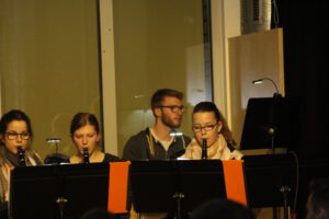 Vier Personen spielen verschiedene Blasinstrumente auf einer Bühne.
