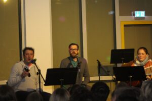 Drei Musiker stehen auf der Bühne und singen in ein Mikrofon.