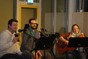 Drei Personen sitzen auf einer Bühne, singen und spielen Instrumente.
