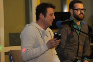 Zwei Männer sitzen bei einem Event, einer hält ein Mikrofon und eine Brille.