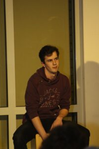 Junger Mann sitzt auf einem Hocker vor einem Fenster und schaut nachdenklich.