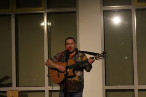 Mann spielt Gitarre und singt auf einer Bühne vor einem Fenster.