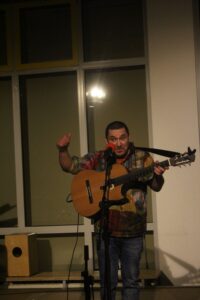 Mann mit Gitarre steht auf einer Bühne und spricht ins Mikrofon.