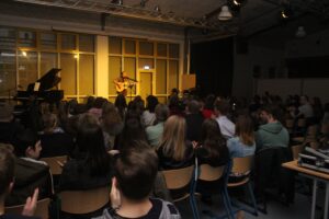 Ein Künstler spielt Gitarre vor einem Publikum in einem Saal.