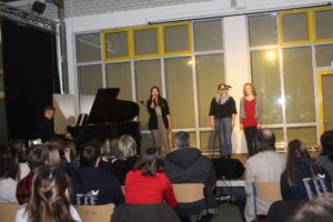 Schülerinnen singen auf der Bühne, Klavierbegleitung im Hintergrund. Publikum sitzt.