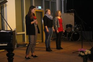 Drei Schülerinnen stehen auf einer Bühne und singen. Im Hintergrund sind Musikinstrumente.