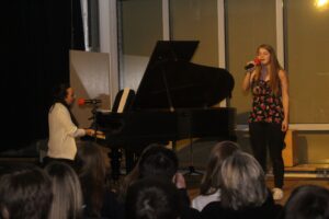 Schülerin singt am Mikrofon, Pianistin begleitet am Klavier. Publikum schaut zu.