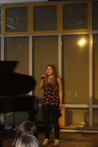 Schülerin singt vor einem Klavier, Publikum im Hintergrund.