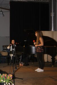Zwei Schülerinnen singen vor einem Klavier auf der Bühne.