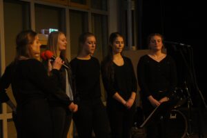 Fünf Frauen stehen auf einer Bühne und singen, eine spricht ins Mikrofon.