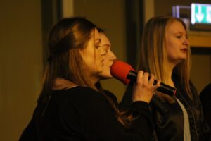 Drei Frauen singen auf einer Bühne, eine hält ein Mikrofon.