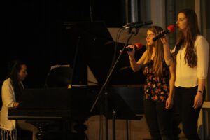 Eine Sängerin mit Mikrofon und eine Pianistin auf einer Bühne.