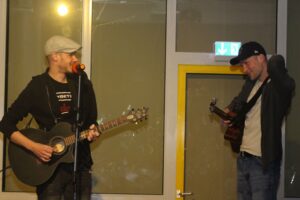 Zwei Männer mit Gitarren stehen auf einer Bühne und spielen Musik.