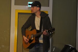 Mann spielt Gitarre in einem Raum mit gelb umrahmtem Fenster.