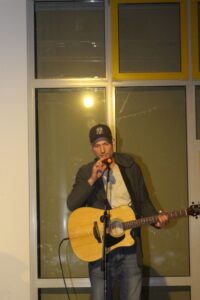 Mann mit Gitarre steht vor einem Fenster und singt ins Mikrofon.