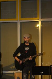 Ein Sänger mit Gitarre steht vor einem Fenster und singt ins Mikrofon.