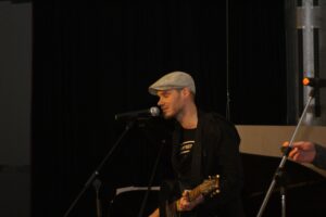 Musiker spielt Gitarre und singt vor Publikum auf einer Bühne.