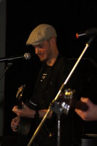 Mann mit Schirmmütze spielt Gitarre und singt in ein Mikrofon.