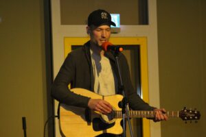Junger Mann mit Gitarre steht am Mikrofon und singt auf einer Bühne.