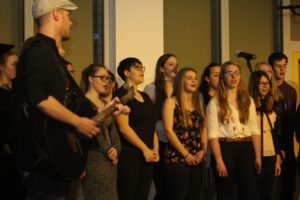 Chor singt auf einer Bühne, Gitarrist spielt im Vordergrund.