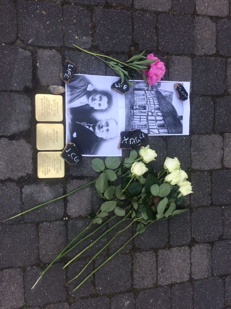 Blumen und Gedenksteine liegen auf einem Pflastersteinboden neben einem Foto.