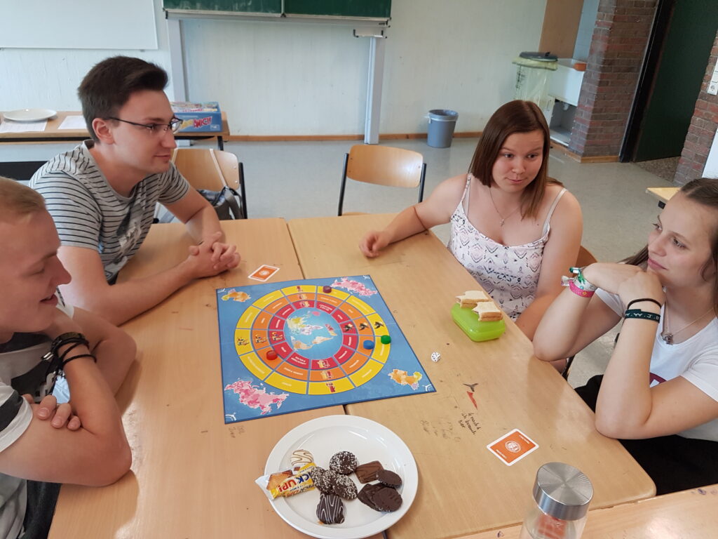 Schüler spielen ein Brettspiel an einem Tisch, Snacks sind ebenfalls vorhanden.