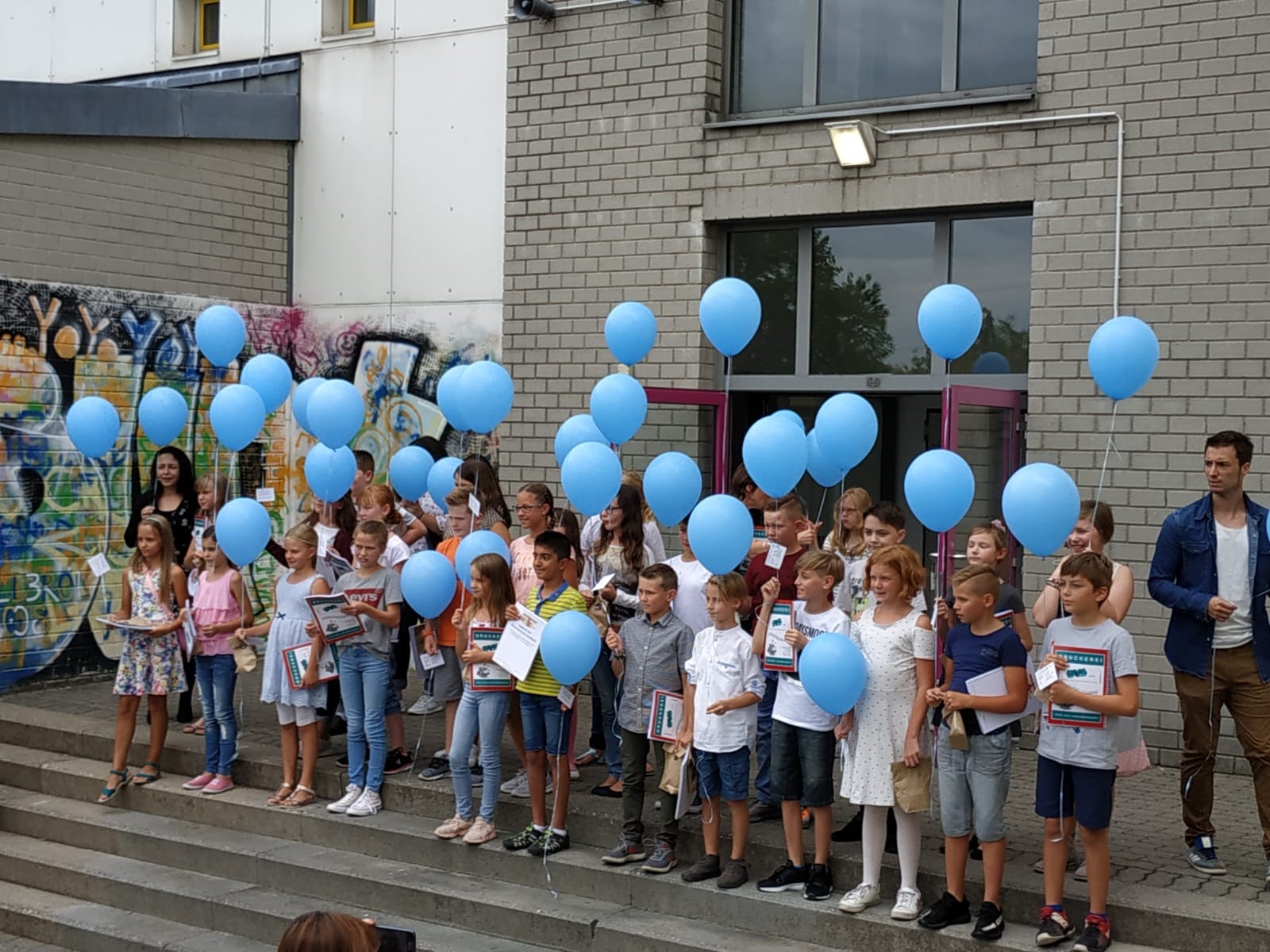 Schüler stehen mit blauen Ballons auf Treppen vor einer Schule.