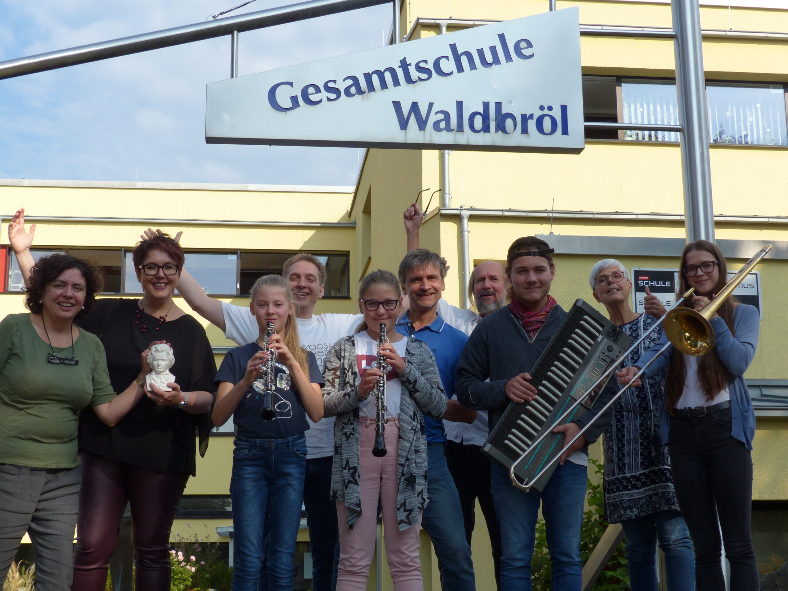 Gruppe von Menschen mit Musikinstrumenten vor dem Schild der Gesamtschule Waldbröl.
