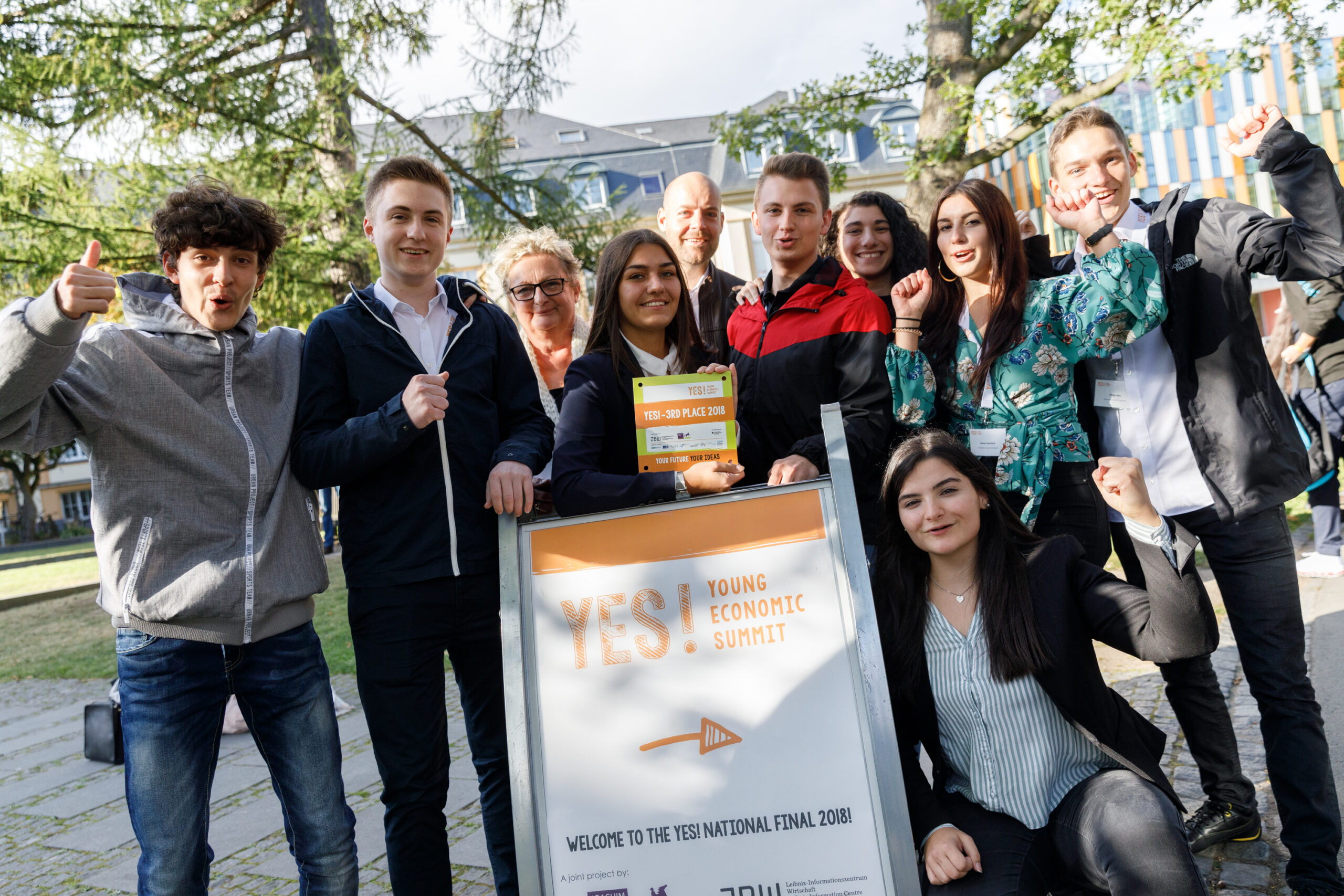 Gruppe von Schülerinnen und Schülern lächelt vor einem Plakat und zeigt Daumen hoch.