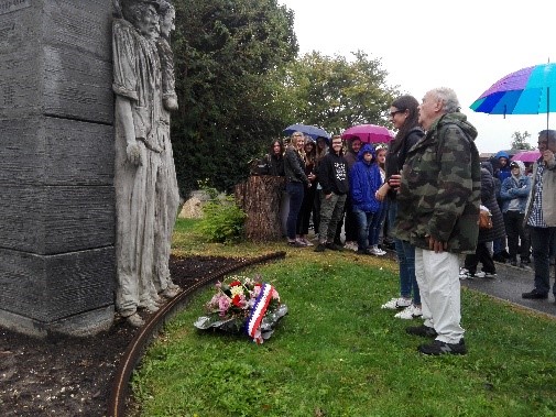 Besucher stehen vor einer Statue, Blumenkranz liegt auf dem Rasen. Es regnet.