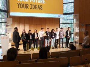 Gruppenvorstellung von Jugendlichen auf einer Bühne mit Präsentation im Hintergrund.
