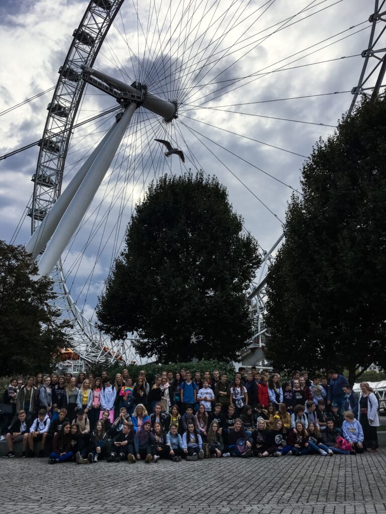 Gruppenselfie vor einem großen Riesenrad unter bewölktem Himmel.