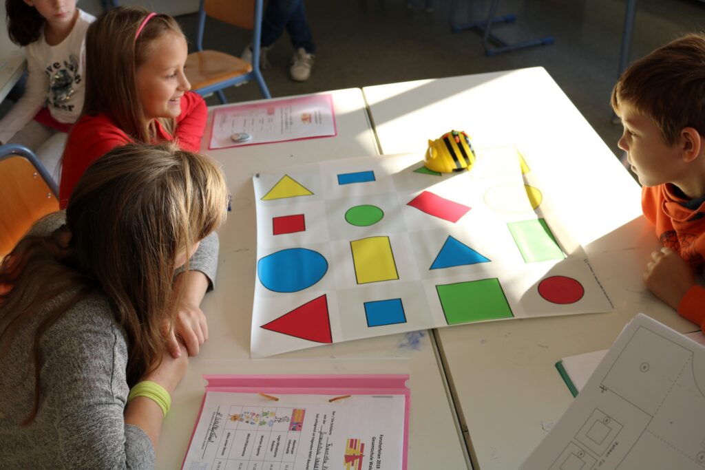 Kinder sitzen am Tisch und betrachten ein Plakat mit verschiedenen Formen in bunten Farben.