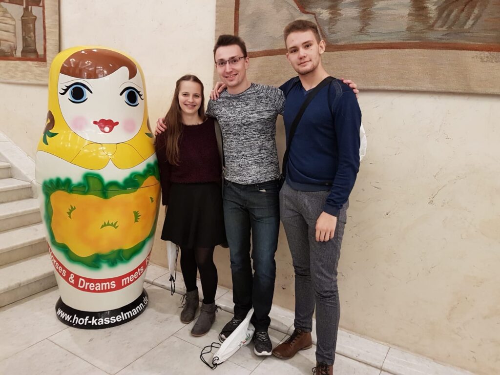 Drei Personen stehen neben einer bunten Matrjoschka-Puppe in einem Gebäude.