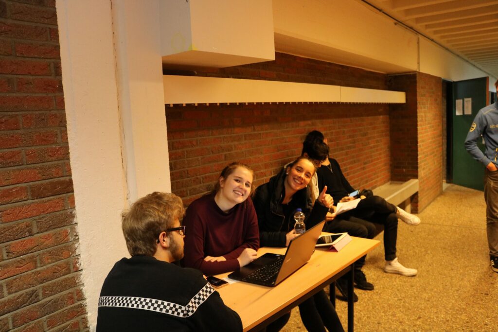 Schüler sitzen an einem Tisch in einem Flur und arbeiten an Laptops.