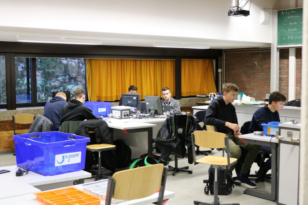 Schüler arbeiten an Tischen im Informatikraum. Computer und Materialien sind sichtbar.