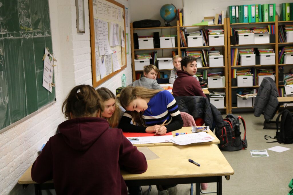 Schüler arbeiten in einem Klassenraum an Aufgaben. Bücherregale im Hintergrund.