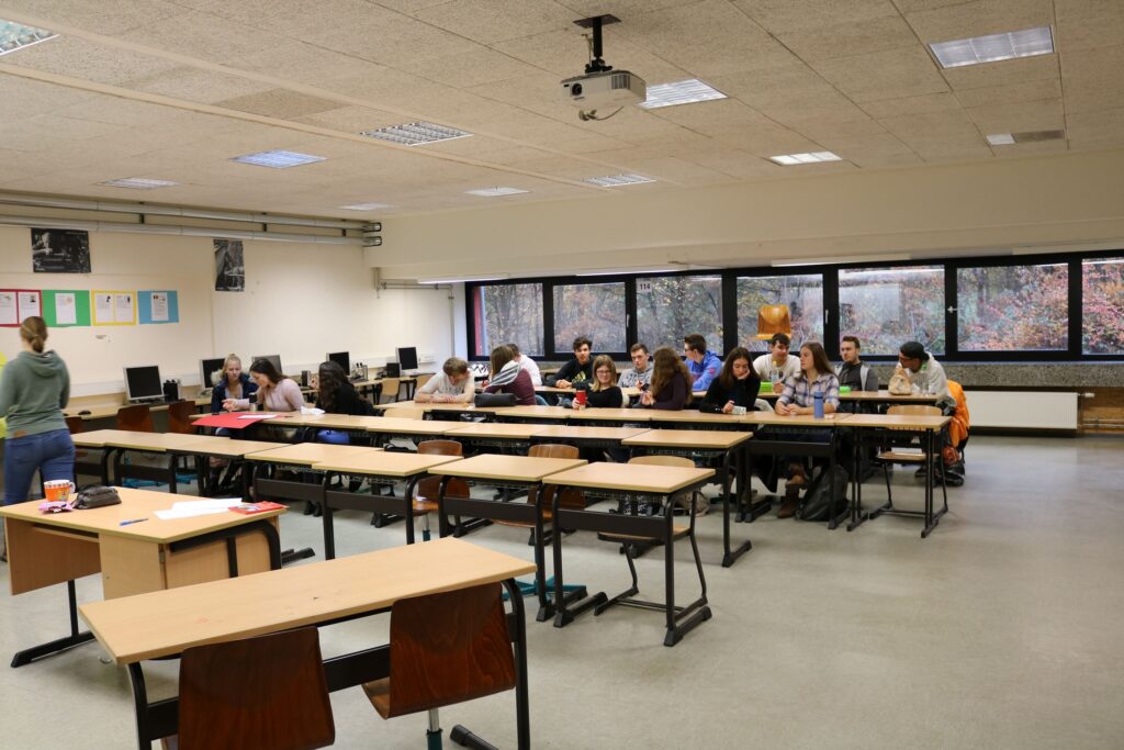 Klassenraum mit Schülern an Tischen, Lehrer spricht vorne.