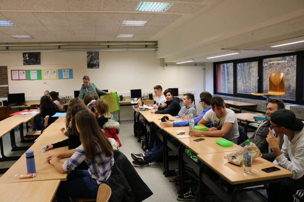 Eine Gruppe von Schülern sitzt an Tischen in einem Klassenraum. Ein Lehrer steht vorne.