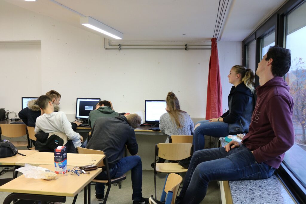 Schüler sitzen in einem Klassenraum vor Computern und kommunizieren miteinander.