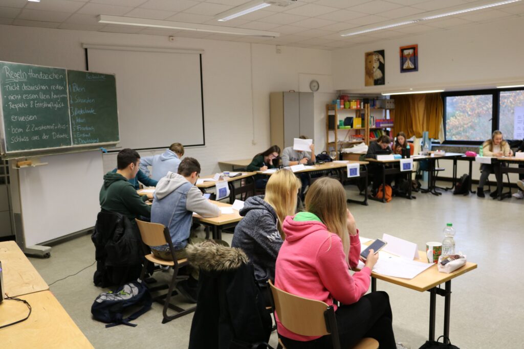 Schüler arbeiten an Tischen in einem Klassenraum mit Tafel.