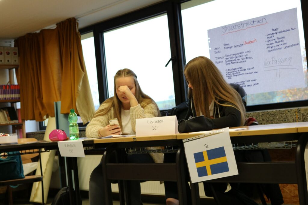 Zwei Schülerinnen sitzen an einem Tisch mit Ländernamen und Flaggen.