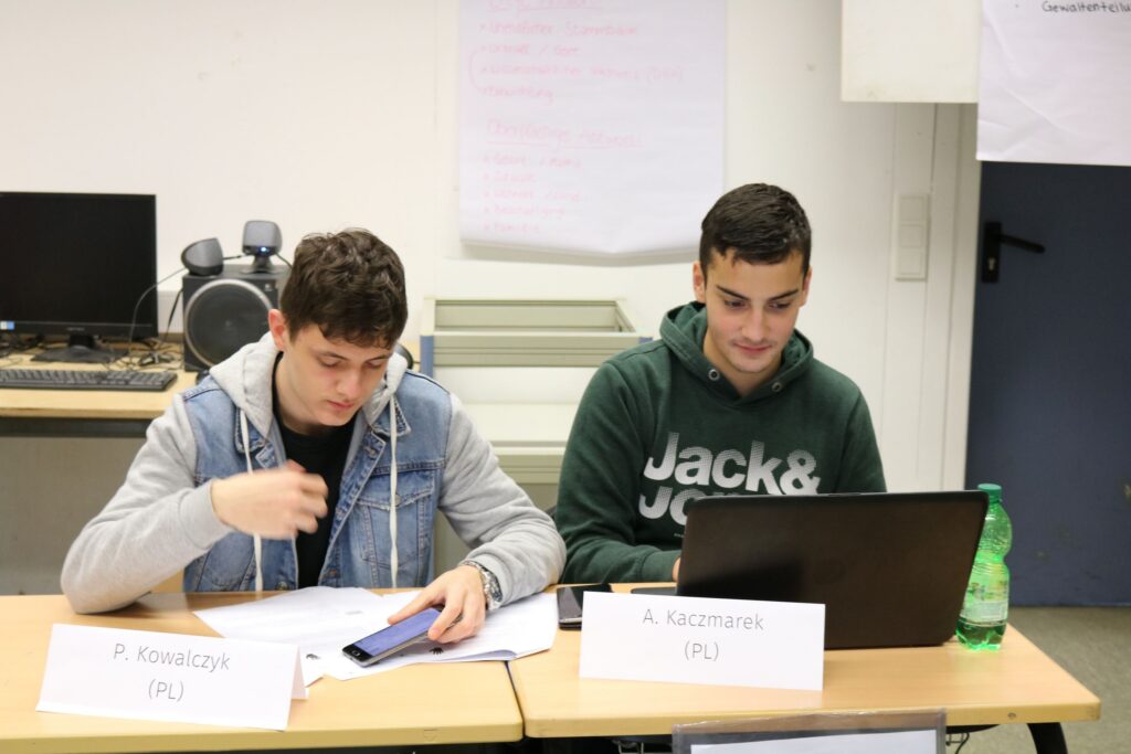Zwei Schüler sitzen an einem Tisch mit Laptop und Unterlagen.